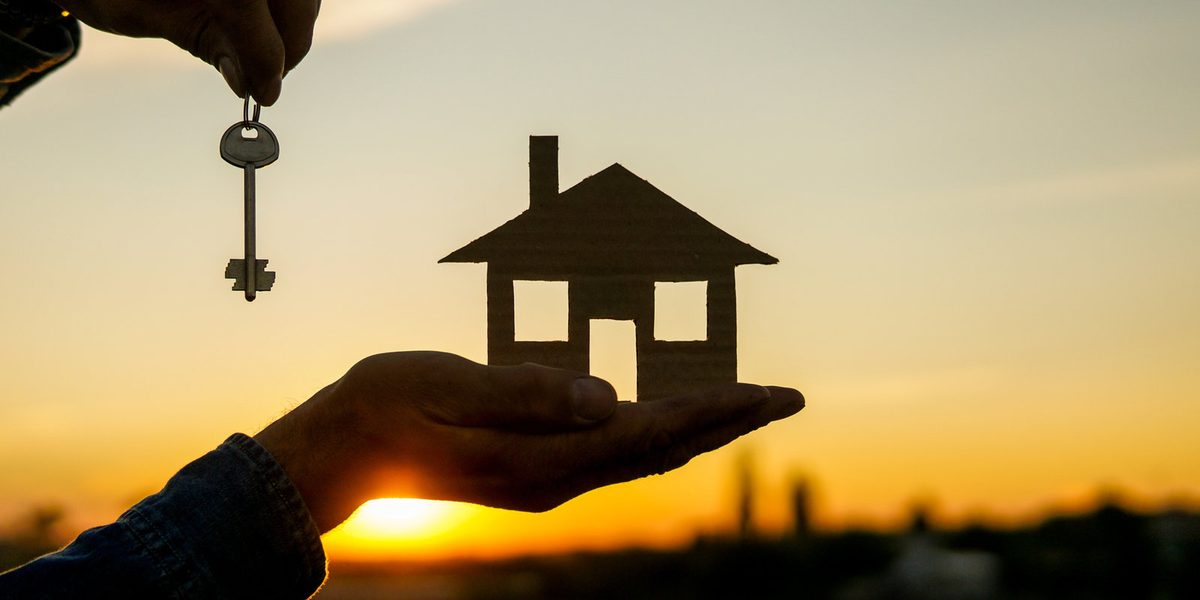 A hand holding a small house cut-out and a key against a sunset — building a stronger community.